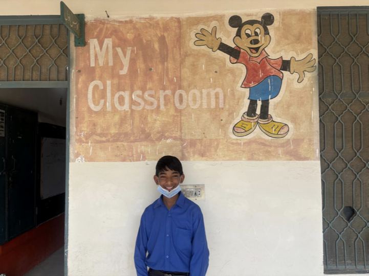 A student smiling in front of his classroom mural at a Punjab government school